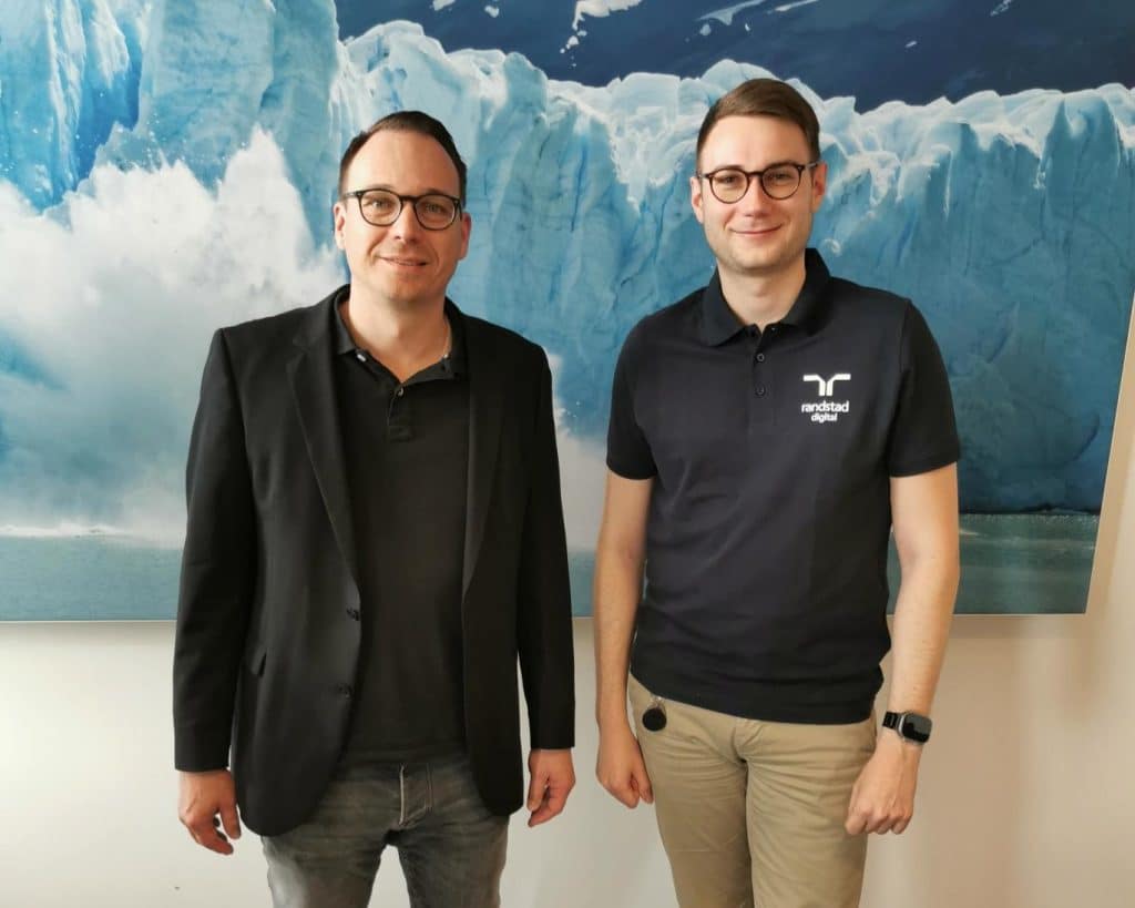 Zwei Männer stehen lächelnd vor einem großen Bild eines eisigen Gletschers. Der Mann links trägt eine schwarze Jacke über einem dunklen Shirt und Jeans, während der Mann rechts ein Polohemd mit dem Logo von 'Randstad Digital' und eine helle Hose trägt. Beide haben eine entspannte Haltung und schauen direkt in die Kamera.