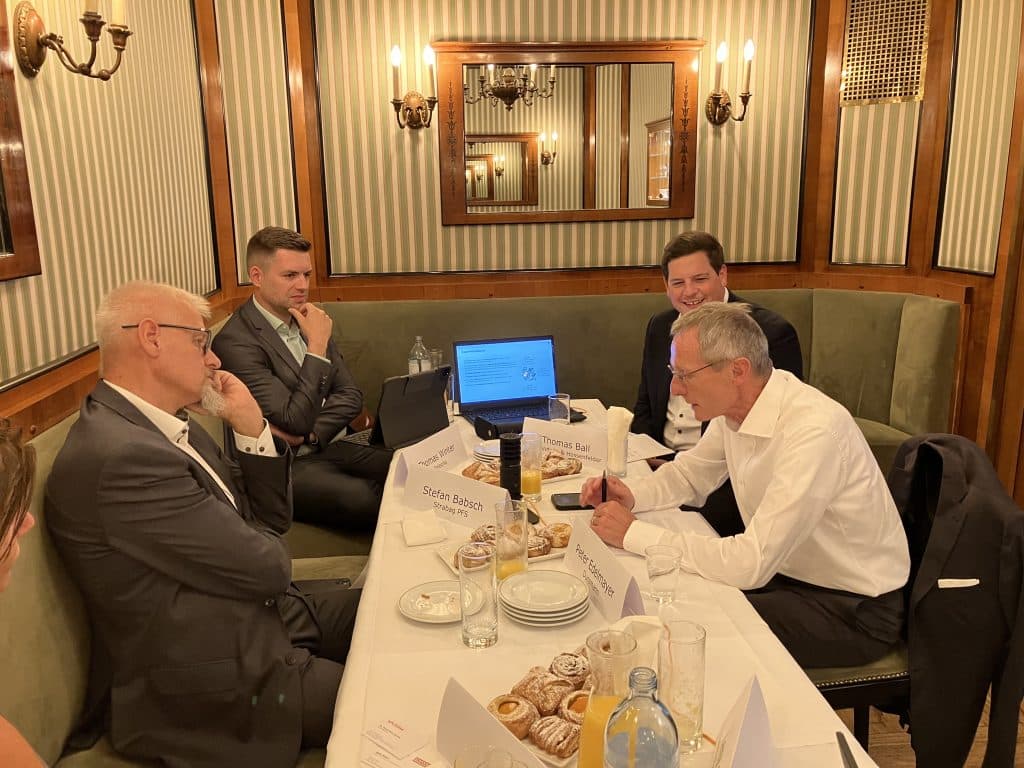 Produktiver Austausch in gemütlicher Atmosphäre: In einem stilvollen Restaurant setzen sich vier Geschäftsleute zu einer intensiven Diskussion zusammen. Mit Notizen, Laptops und Namensschildern vor sich, tauschen sie Ideen aus und besprechen relevante Themen. Die entspannte, aber konzentrierte Stimmung zeigt die Bedeutung des Treffens – ein inspirierender Moment für strategische Entscheidungen und erfolgreiche Zusammenarbeit.