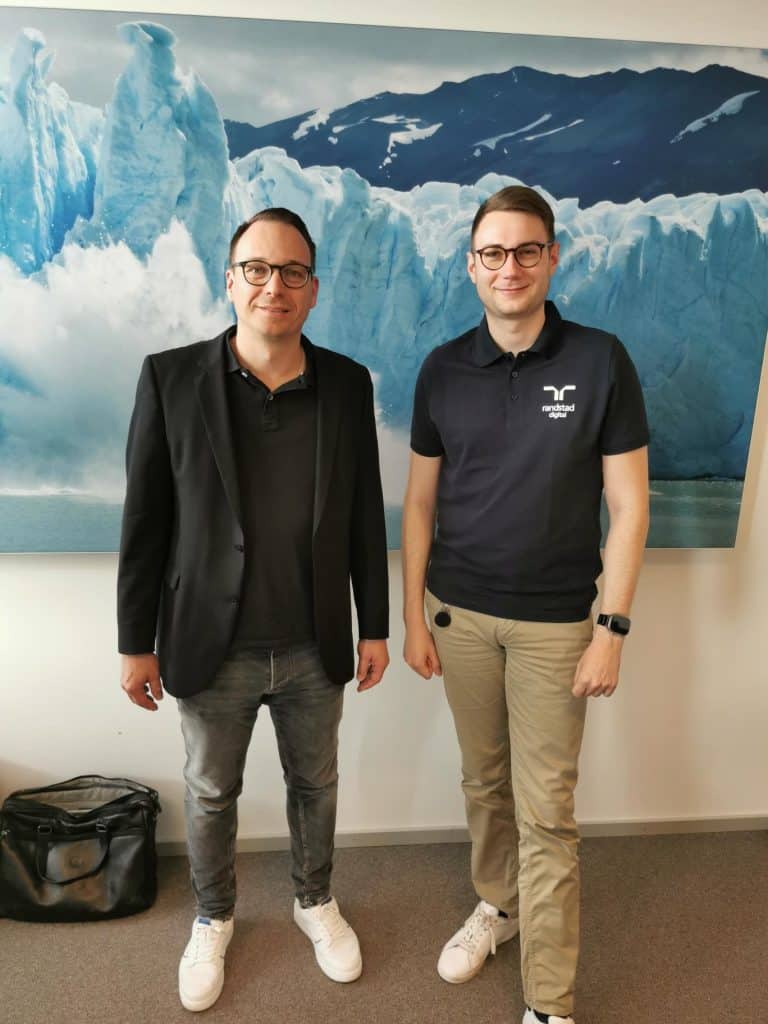 Zwei Männer stehen lächelnd vor einem großen Bild eines eisigen Gletschers. Der Mann links trägt eine schwarze Jacke über einem dunklen Shirt und Jeans, während der Mann rechts ein Polohemd mit dem Logo von 'Randstad Digital' und eine helle Hose trägt. Beide haben eine entspannte Haltung und schauen direkt in die Kamera.
