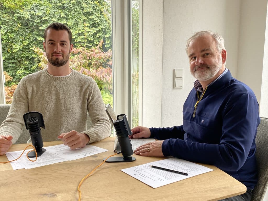 Zwei Männer sitzen an einem Holztisch in einem hellen Raum mit großen Fenstern, durch die eine grüne Gartenlandschaft sichtbar ist. Vor ihnen liegen Dokumente und Mikrofone auf Stativen, was auf eine Podcast- oder Interviewaufnahme hindeutet. Der jüngere Mann auf der linken Seite trägt einen hellen Strickpullover und hat kurze dunkle Haare, während der ältere Mann auf der rechten Seite einen dunkelblauen Pullover mit einem grauen Bart trägt. Beide blicken freundlich in die Kamera.