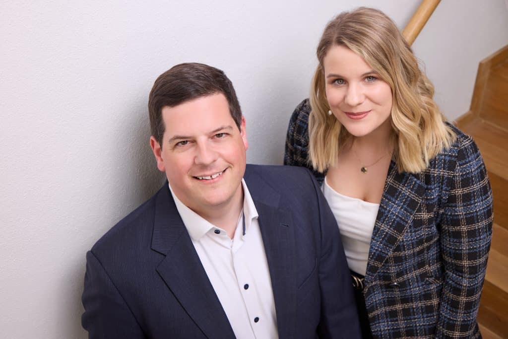 Ein professionelles Business-Porträt eines Mannes und einer Frau in eleganter Kleidung. Der Mann trägt einen dunkelblauen Anzug mit weißem Hemd und lächelt freundlich in die Kamera. Die Frau steht leicht hinter ihm, trägt eine stilvolle karierte Blazerjacke mit weißem Top und hat blonde, wellige Haare. Beide blicken freundlich und selbstbewusst in die Kamera. Der Hintergrund ist neutral mit einer weißen Wand und einer Holztreppe, was eine warme und professionelle Atmosphäre schafft.