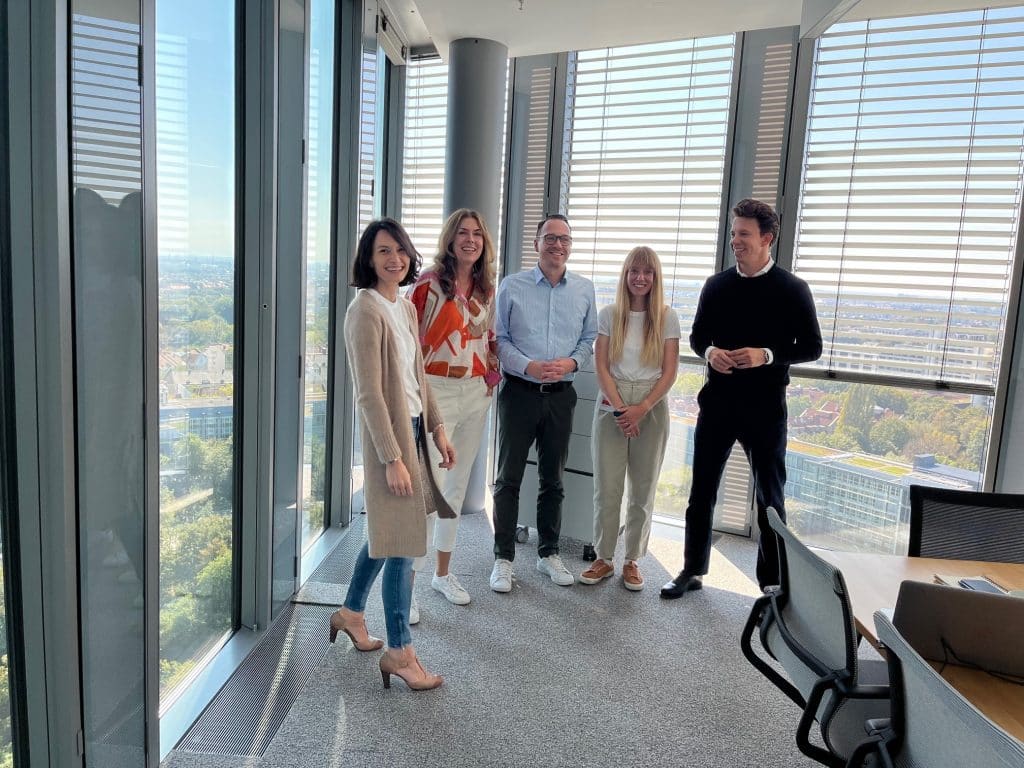 Eine Gruppe von fünf Personen steht in einem modernen Büro mit großen Fenstern und Blick auf eine Stadtlandschaft. Sie lächeln und scheinen sich in einem entspannten Gespräch zu befinden. Die Atmosphäre wirkt professionell und freundlich, mit einer Mischung aus formeller und legerer Kleidung. Das Büro ist hell und einladend, mit einem Konferenztisch und Stühlen im Vordergrund.