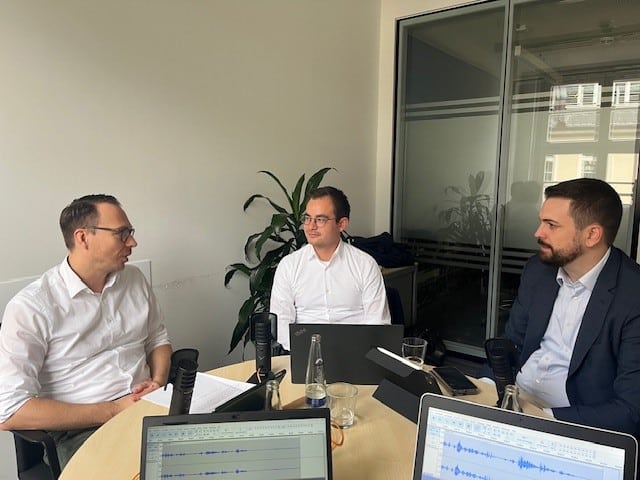 Drei Männer sitzen an einem runden Tisch in einem modernen, hellen Büro und führen eine Diskussion. Vor ihnen stehen Mikrofone, Laptops und Notizen, während eine Tonaufnahme-Software auf den Bildschirmen sichtbar ist. Zwei der Männer tragen weiße Hemden, einer einen dunklen Anzug. Die Atmosphäre wirkt professionell und konzentriert.
