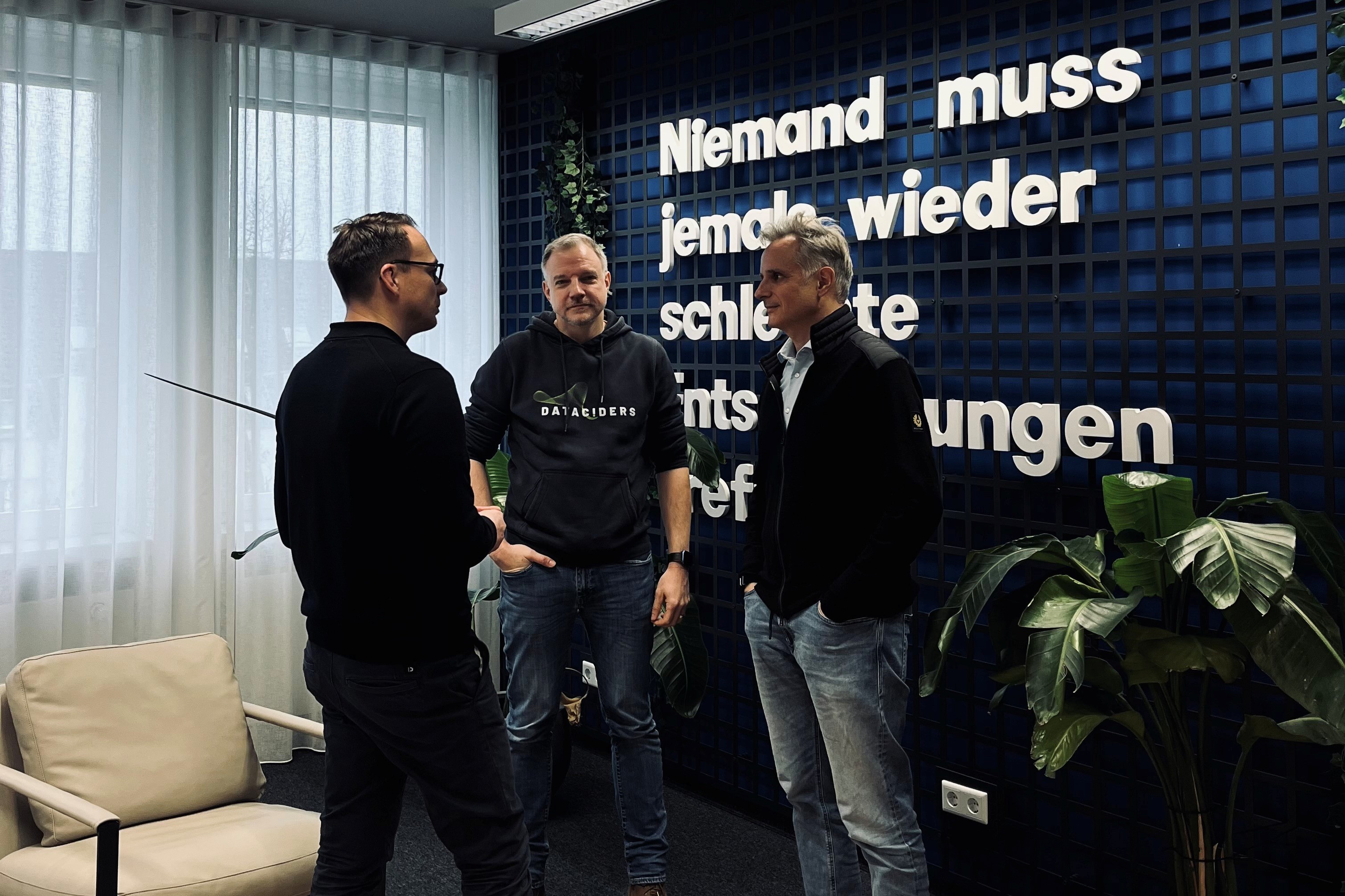 Mario Zillmann, Gero Presser und Christian Schneider im Gespräch in einem Büroraum. Im Hintergrund zieht man den Schriftzug "Niemand muss jemals wieder schlechre Entscheidungen treffen."