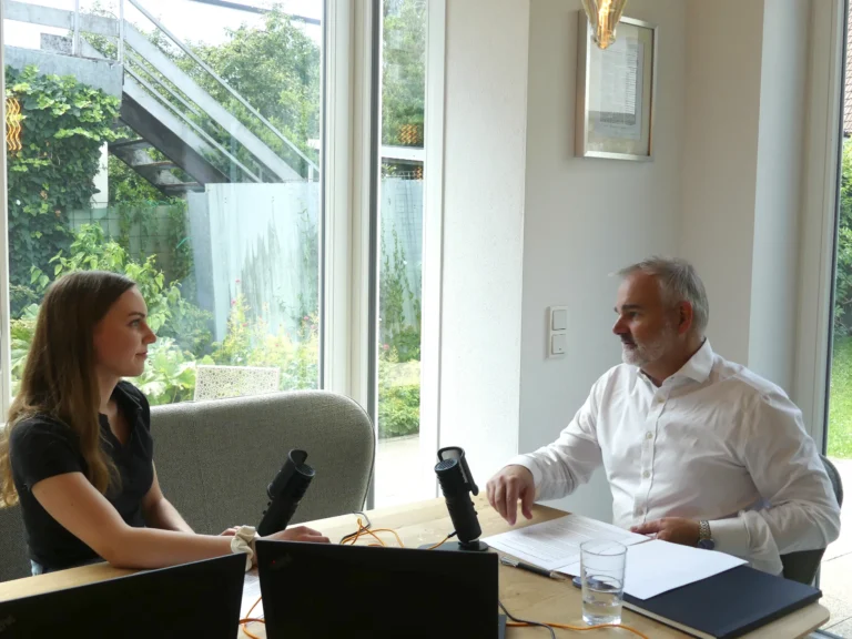 Zwei Personen sitzen sich an einem Holztisch gegenüber und führen ein Interview. Auf dem Tisch stehen zwei schwarze Podcast-Mikrofone sowie ein Laptop und ein Wasserglas. Die Person links trägt ein schwarzes Oberteil und hört aufmerksam zu; die Person rechts trägt ein weißes Hemd und spricht. Im Hintergrund ist eine große Fensterfront mit Blick auf einen grünen Garten sowie eine Außentreppe zu sehen.