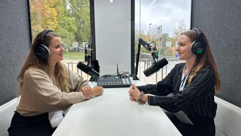 Zwei Frauen sitzen sich in einer Podcast-Aufnahmekabine gegenüber und lächeln sich an. Beide tragen Kopfhörer, vor ihnen stehen Studiomikrofone an Schwenkarmen und ein Mischpult auf einem weißen Tisch.